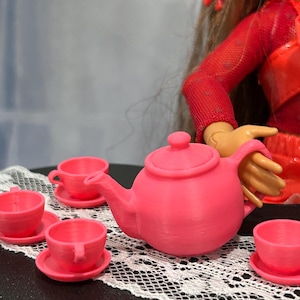 Peut inclure: Un service à thé rose miniature, comprenant une théière, des soucoupes et des tasses, est disposé sur un napperon en dentelle blanche. L'ensemble est en plastique et est conçu pour les poupées ou autres jouets. La théière a un couvercle et une anse.