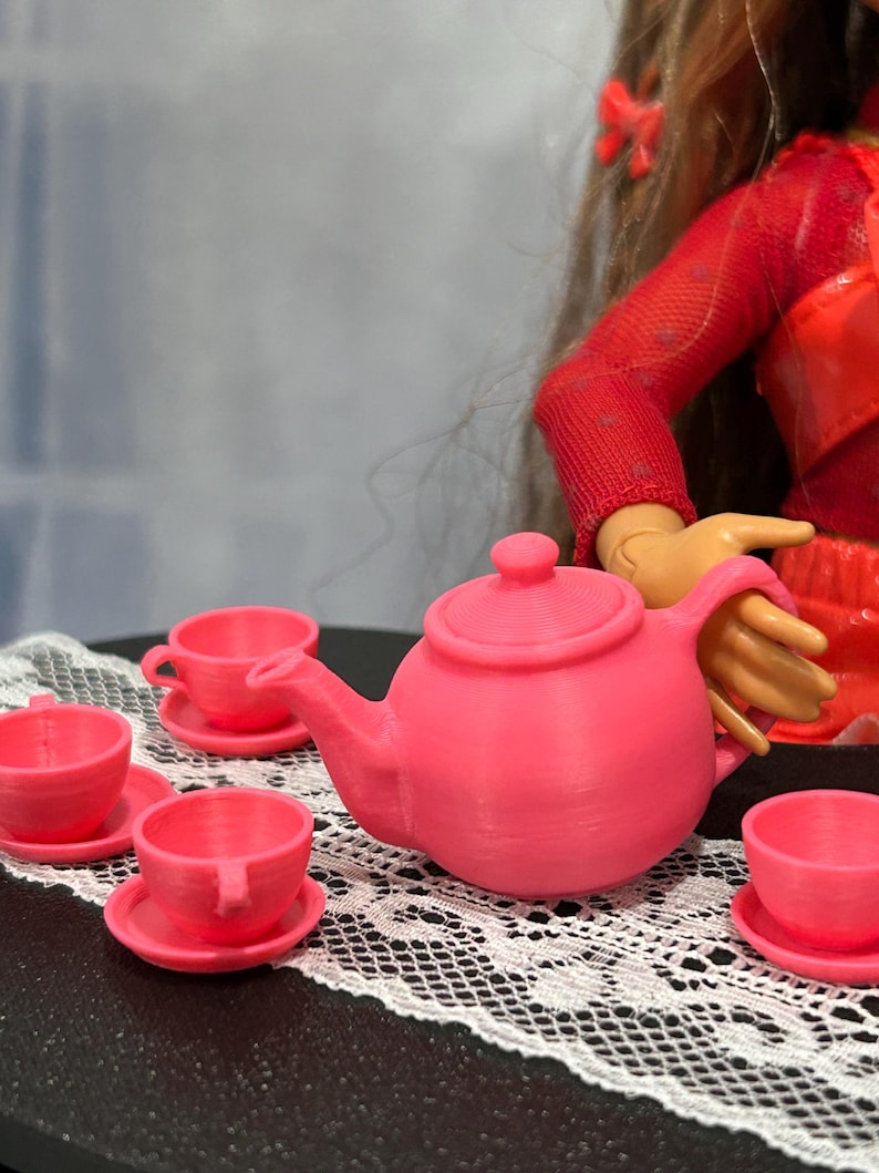 May include: A miniature pink tea set, including a teapot, saucers, and teacups, is arranged on a white lace doily. The set is a vibrant pink color. The scene suggests a doll-sized tea party.