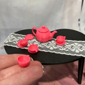 May include: A miniature pink tea set, including a teapot, saucers, and teacups, arranged on a small black table with a white lace runner. The set is made of a solid pink material. A hand holds a teacup in the foreground.