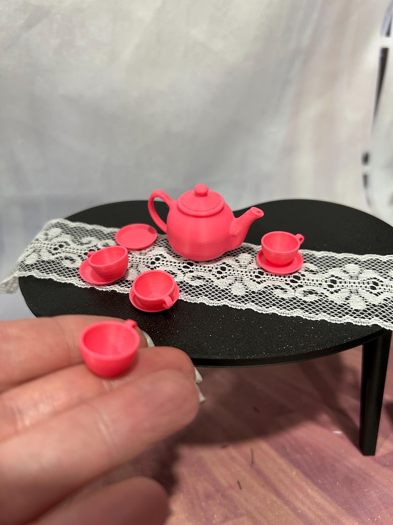 May include: A miniature pink tea set, including a teapot, saucers, and teacups, arranged on a small black table with a white lace runner. The set is made of a solid pink material. A hand holds a teacup in the foreground.