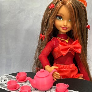 May include: A doll with long brown hair and red accents is seated at a table set for tea. The doll wears a red outfit with a large bow. A pink teapot and matching teacups are arranged on a table with a white lace runner, creating a miniature tea party scene.