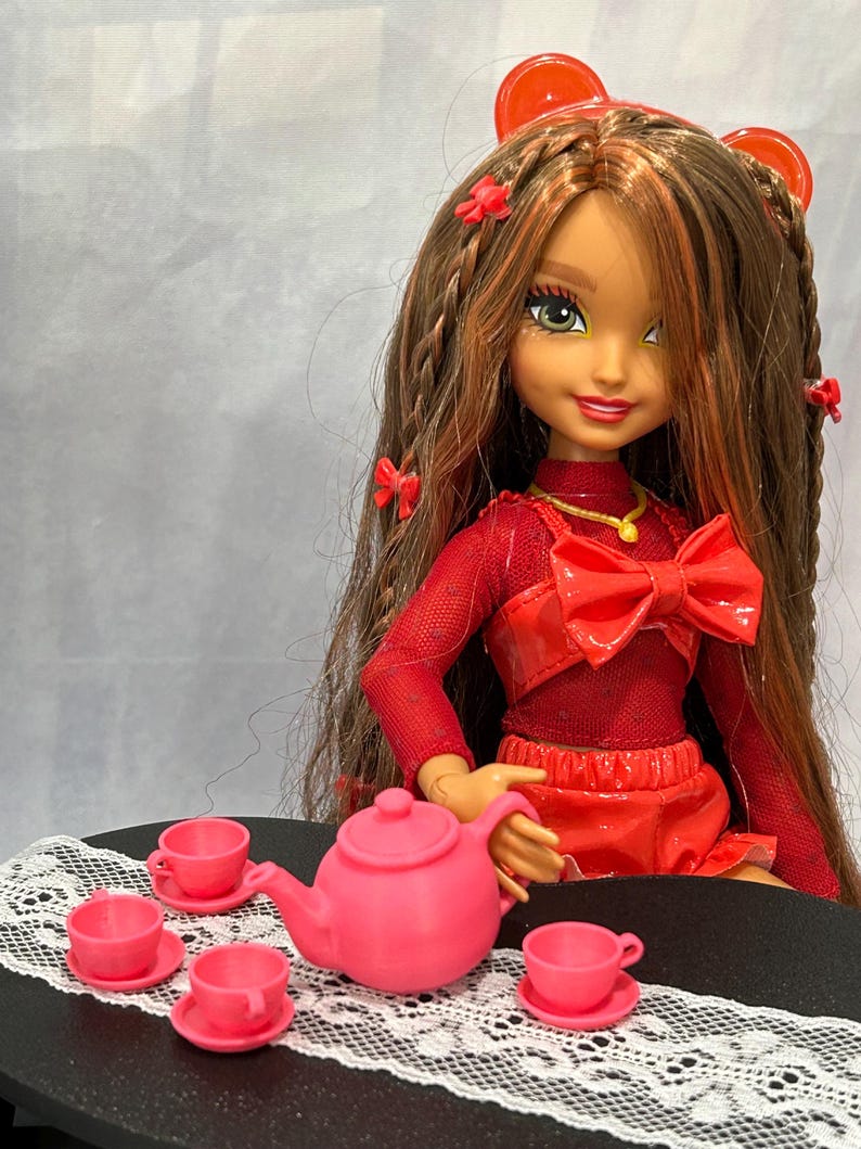 May include: A doll with long brown hair and red accents is seated at a table set for tea. The doll wears a red outfit with a large bow. A pink teapot and matching teacups are arranged on a table with a white lace runner, creating a miniature tea party scene.