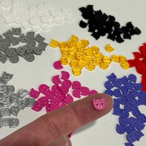 Peut inclure: Une collection de petits boutons en forme de chat de différentes couleurs, notamment blanc, noir, gris, jaune, rouge, rose, argent et violet. Chaque bouton possède deux trous pour la couture. Un bouton rose en forme de chat est présenté.
