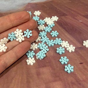 May include: A collection of small, flat, two-hole buttons shaped like snowflakes. The buttons are colored in alternating shades of solid white and light pastel blue. Some buttons are held in a hand, while others are scattered across the surface.