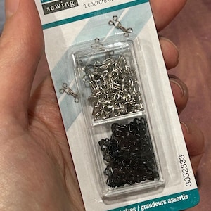 May include: A pack of 30 assorted size silver and black sew-on hooks and eyes. The pack is labelled "UNIQUE sewing 30 agrafes à courdre et œillets 30 hooks & sew on assorted sizes/grandeurs assortis 3032333".