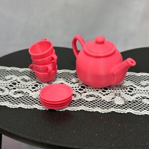 May include: A miniature pink tea set, including a teapot, three teacups, and a saucer. The teapot has a handle and a spout. The teacups are stacked. The set is displayed on a black surface with a white lace runner.