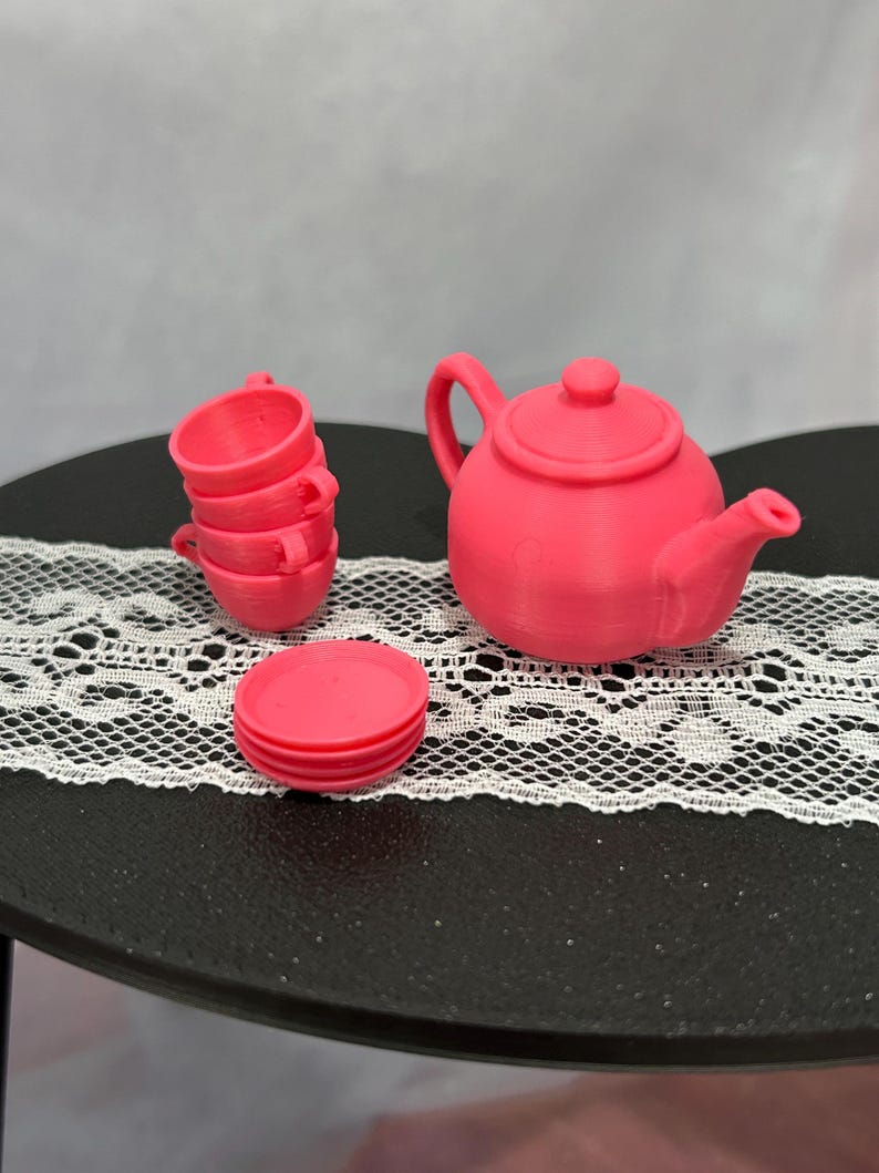 May include: A miniature pink tea set, including a teapot, three teacups, and a saucer. The teapot has a handle and a spout. The teacups are stacked. The set is displayed on a black surface with a white lace runner.