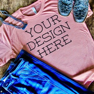 May include: A pink t-shirt with the text "YOUR DESIGN HERE" printed on the front. The shirt is lying on a brown surface with a pair of blue jeans and a necklace.