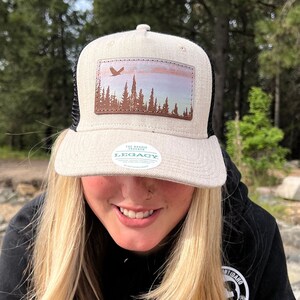 May include: A beige and black trucker hat with a brown leather patch featuring a silhouette of a forest and a bird flying over a sunset. The hat has a green sticker with the text "The Roadie Traveler Legacy" on the front.