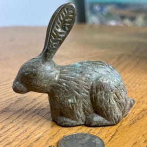 May include: A small, bronze rabbit figurine with a textured surface. The rabbit is sitting with its ears up and its head tilted to the side.