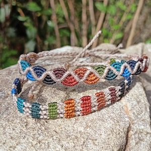 Könnte beinhalten: Zwei bunte Freundschaftsarmbänder aus geflochtener Schnur in den Farben Braun, Rot, Grün, Blau und Schwarz. Die Armbänder liegen auf einem grauen Stein.