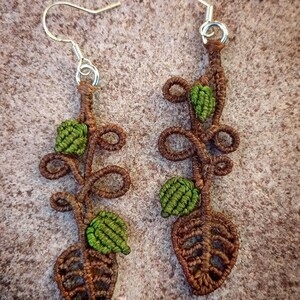 Peut inclure: Une paire de boucles d'oreilles en forme de feuille de couleur marron et verte avec des crochets argentés. Les boucles d'oreilles sont en macramé et présentent un délicat motif de vigne avec de petites feuilles vertes.