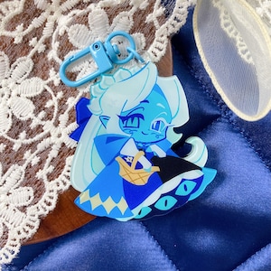 Op de afbeelding: Een doorzichtige acryl sleutelhanger met een gestileerde illustratie van een lichtblauw personage met lang wit haar, gekleed in een blauwe jurk en cape, met een klein beige mandje, bevestigd aan een lichtblauwe plastic karabijnhaak.