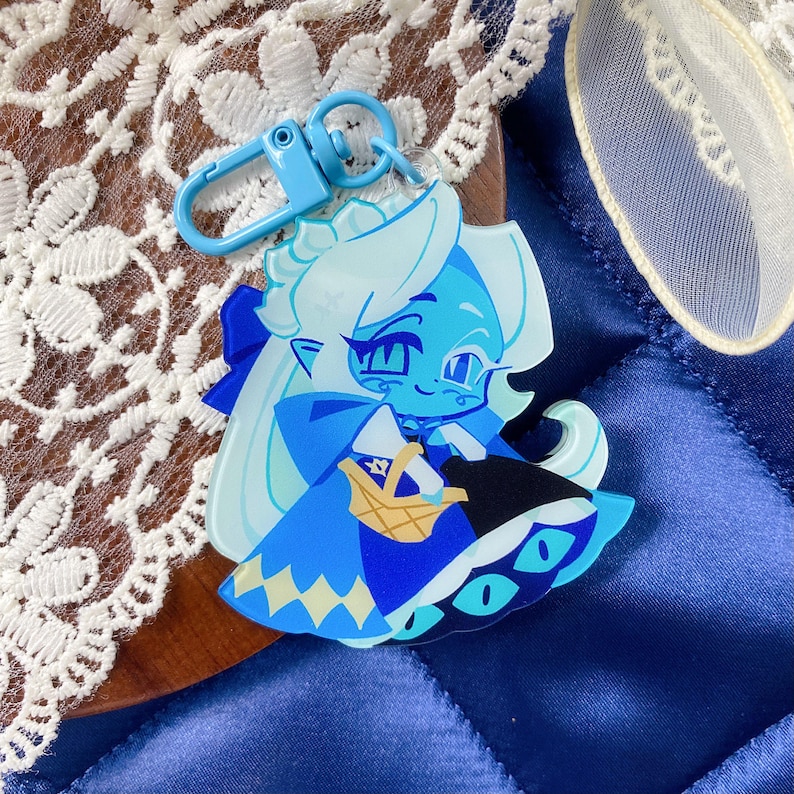 Op de afbeelding: Een doorzichtige acryl sleutelhanger met een gestileerde illustratie van een lichtblauw personage met lang wit haar, gekleed in een blauwe jurk en cape, met een klein beige mandje, bevestigd aan een lichtblauwe plastic karabijnhaak.