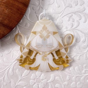 Op de afbeelding: Een doorschijnende acryl hanger met een gestileerd figuur met wit haar en een goudkleurig hoofddeksel. Het figuur draagt een vloeiend wit en goud gewaad met geometrische patronen en houdt een klein gouden voorwerp vast, bekroond met een witte plastic ring.