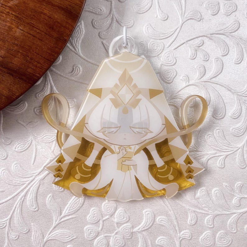 Op de afbeelding: Een doorschijnende acryl hanger met een gestileerd figuur met wit haar en een goudkleurig hoofddeksel. Het figuur draagt een vloeiend wit en goud gewaad met geometrische patronen en houdt een klein gouden voorwerp vast, bekroond met een witte plastic ring.