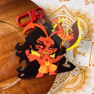 Op de afbeelding: Een sleutelhanger van een roodharig personage met zwart haar, scherpe tanden en gloeiende ogen, gekleed in een geel en oranje harnas. Het personage houdt een grote bijl met een geel blad vast. Een rode plastic karabijnhaak zit aan de bovenkant.