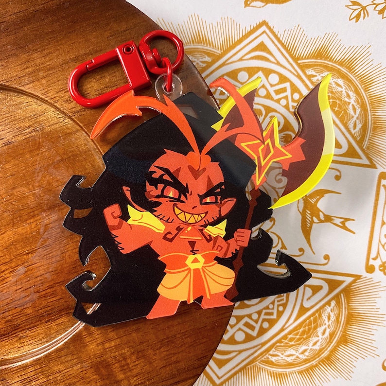 Op de afbeelding: Een sleutelhanger van een roodharig personage met zwart haar, scherpe tanden en gloeiende ogen, gekleed in een geel en oranje harnas. Het personage houdt een grote bijl met een geel blad vast. Een rode plastic karabijnhaak zit aan de bovenkant.