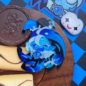Op de afbeelding: Een blauwe en zwarte acryl hanger met een gestileerd personage met een narrenkap, die een staf met een oogvormige edelsteen vasthoudt. Het personage heeft een lichtblauwe huid, donkerblauw haar en een brede grijns. Een kleine witte hartvormige pin.