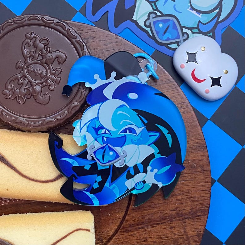Op de afbeelding: Een blauwe en zwarte acryl hanger met een gestileerd personage met een narrenkap, die een staf met een oogvormige edelsteen vasthoudt. Het personage heeft een lichtblauwe huid, donkerblauw haar en een brede grijns. Een kleine witte hartvormige pin.