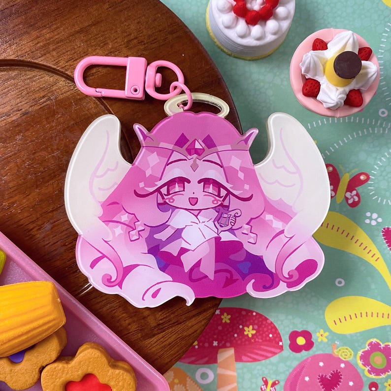 Op de afbeelding: Een roze acryl sleutelhanger met een gestileerd figuur met lang golvend haar, een kroon en vleugels. Het figuur heeft een kleine halo boven het hoofd en houdt een piepkleine harp vast. Inclusief een roze plastic karabijnhaak als bevestiging.