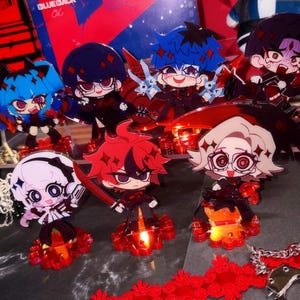 May include: A collection of seven acrylic chibi-style character standees featuring diverse hair colors including blue, black, red, and blonde. Each figure is mounted on a translucent red circular base, with some figures holding weapons like scythes.