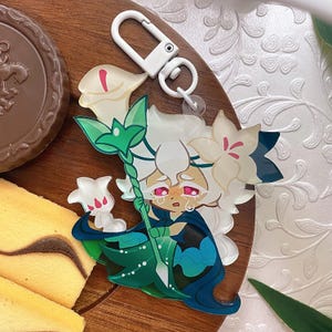 Op de afbeelding: Een acryl sleutelhanger met een cartoonfiguur met wit haar, roze ogen en een betraande uitdrukking. Het figuur draagt een donkerblauwe cape en houdt een groene staf vast die bekroond is met een witte lelie. Een witte plastic sluiting is bevestigd.