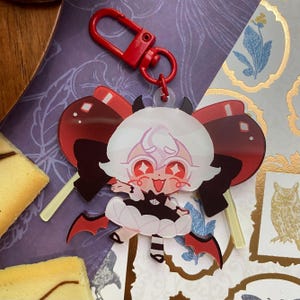 Op de afbeelding: Een doorzichtige acryl sleutelhanger met een cartoonfiguur met wit haar, rode ogen en een zwart-witte outfit met kleine vleermuisvleugels. Het figuur is geplaatst voor een grote rode strik en is bevestigd aan een felrode plastic karabijnhaak.