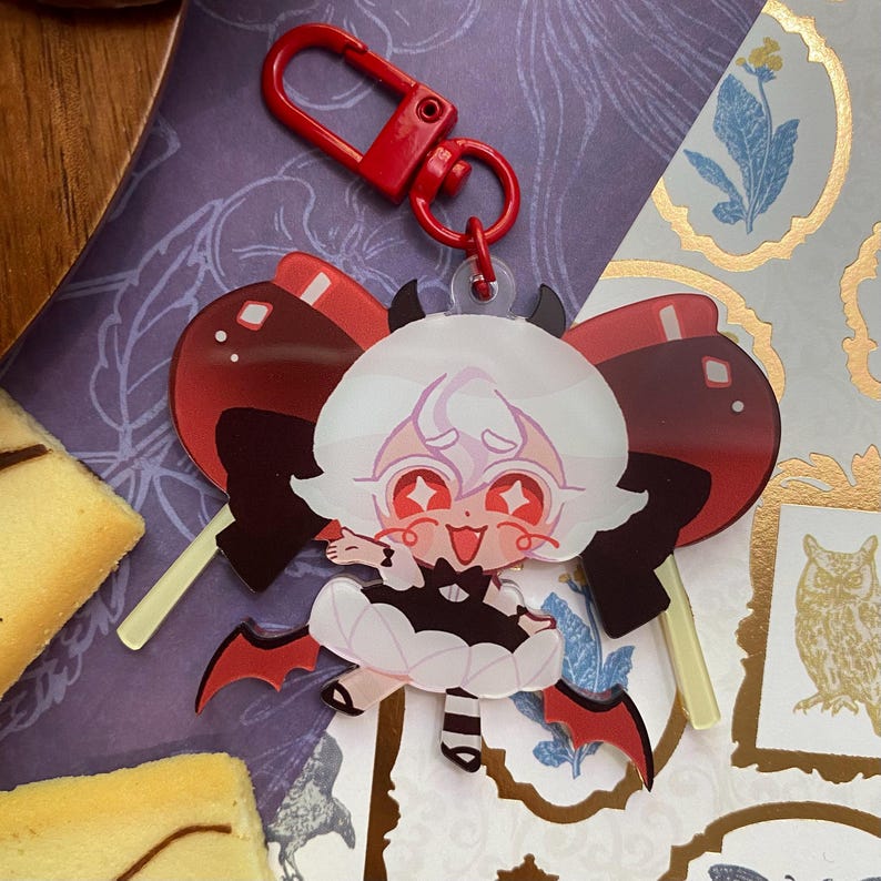 Op de afbeelding: Een doorzichtige acryl sleutelhanger met een cartoonfiguur met wit haar, rode ogen en een zwart-witte outfit met kleine vleermuisvleugels. Het figuur is geplaatst voor een grote rode strik en is bevestigd aan een felrode plastic karabijnhaak.