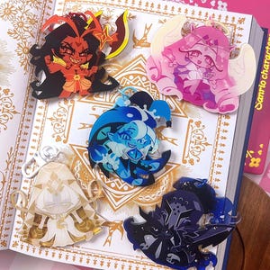 Op de afbeelding: Vijf acryl bedels met diverse ontwerpen: een rode demon met een wapen, een roze gevleugelde figuur, een blauw personage in narrenstijl, een wit geklede figuur met gouden accenten en een donkerblauwe gepantserde ridder met een zwaard.