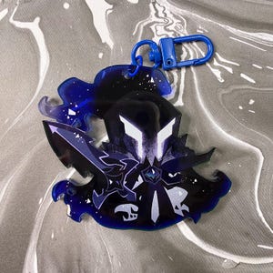 Op de afbeelding: Een donkerblauwe en zwarte acryl sleutelhanger met een gestileerd personage-silhouet met witte hoekige ogen, die een zwaard vasthoudt. Het ontwerp bevat een doorschijnende blauwe rand met witte spikkels en een blauwe plastic karabijnhaak.