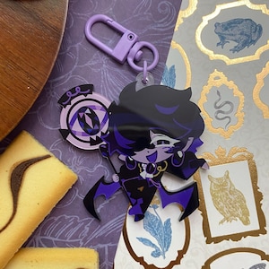 Op de afbeelding: Een paarse en zwarte acryl sleutelhanger met een gestileerd stripfiguur met donker haar, gekleed in een formeel pak met paarse accenten, en een cirkelvormige staf met een paars oogontwerp vasthoudend, bevestigd aan een lichtpaarse plastic clip.