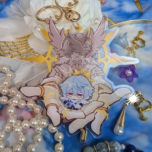 May include: A clear acrylic charm featuring a white-winged angel with gold accents holding a small blue-haired character. The charm is surrounded by a gold halo and stars.