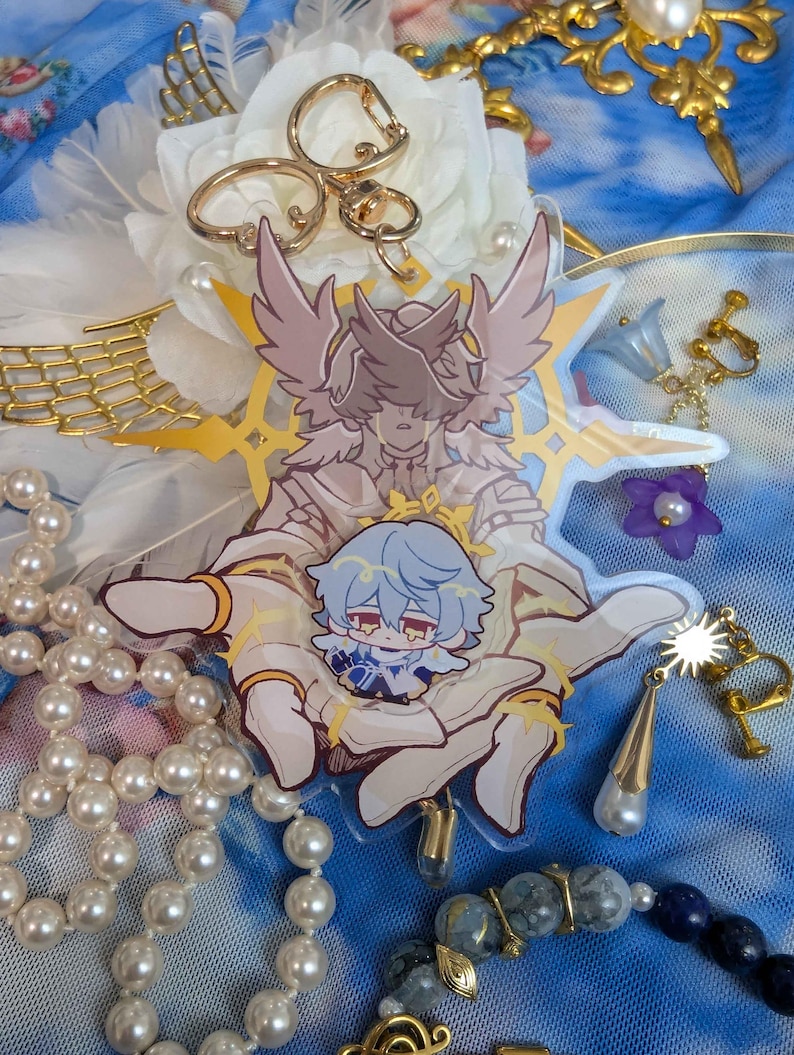 May include: A clear acrylic charm featuring a white-winged angel with gold accents holding a small blue-haired character. The charm is surrounded by a gold halo and stars.