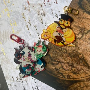 Op de afbeelding: Twee acryl sleutelhangers met gestileerde stripfiguren. De ene toont een personage met een hoge hoed, gele bril en een gouden sleutel, de andere een personage met wit haar, rode accenten en groene hoorns.