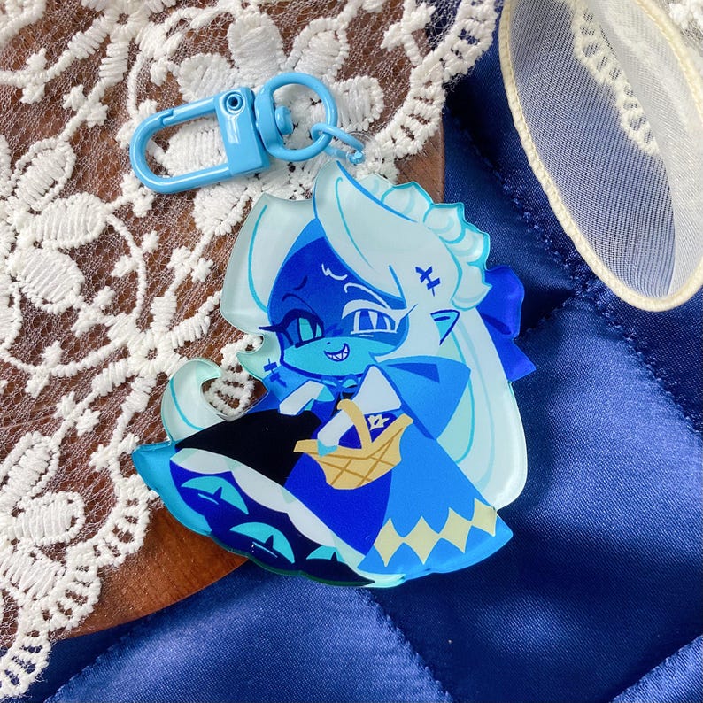 Op de afbeelding: Een lichtblauwe acryl sleutelhanger met een gestileerd personage met lang wit haar, puntige oren en een blauwe outfit. Het personage houdt een klein geel mandje vast. De sleutelhanger is voorzien van een bijpassende lichtblauwe plastic karabijnhaak.