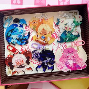 Op de afbeelding: Een verzameling van zes acryl sleutelhangers met diverse anime-stijl figuren in levendige kleuren, waaronder blauw, goud, groen, rood, paars en roze, elk bevestigd aan een bijpassende plastic sluiting, gepresenteerd in een rechthoekige doos.