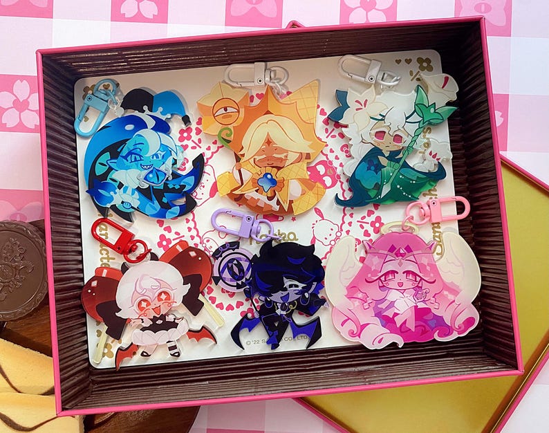 Op de afbeelding: Een verzameling van zes acryl sleutelhangers met diverse anime-stijl figuren in levendige kleuren, waaronder blauw, goud, groen, rood, paars en roze, elk bevestigd aan een bijpassende plastic sluiting, gepresenteerd in een rechthoekige doos.