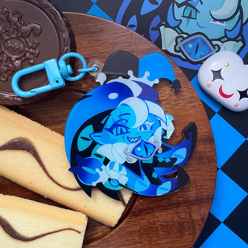 Op de afbeelding: Een blauwe en zwarte acryl sleutelhanger met een gestileerd personage met een narrenkap, scherpe tanden en een blauw oogvormig accessoire. De sleutelhanger heeft een lichtblauwe karabijnhaak en ligt op een houten ondergrond.