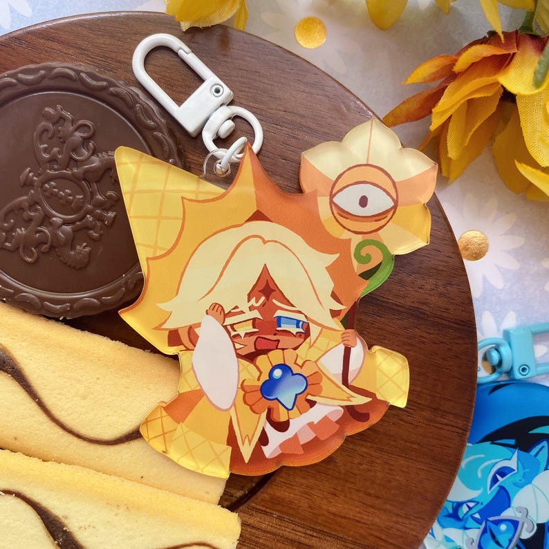 Op de afbeelding: Een acryl sleutelhanger met een cartoonfiguur met blond haar, gekleed in een geel en oranje outfit met een blauw ster-embleem, die een staf vasthoudt met een gele bloem met een oog erin, bevestigd aan een witte plastic karabijnhaak.