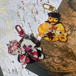 May include: Two acrylic keychains featuring stylized cartoon characters. One keychain has a red clasp and depicts a purple-skinned character with white hair and horns. The other has a gold clasp and shows a yellow-haired character wearing a top hat.