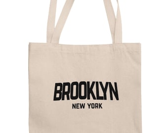 May include: A beige tote bag featuring the word "BROOKLYN" in bold black lettering, with "NEW YORK" underneath. The bag has two beige handles.