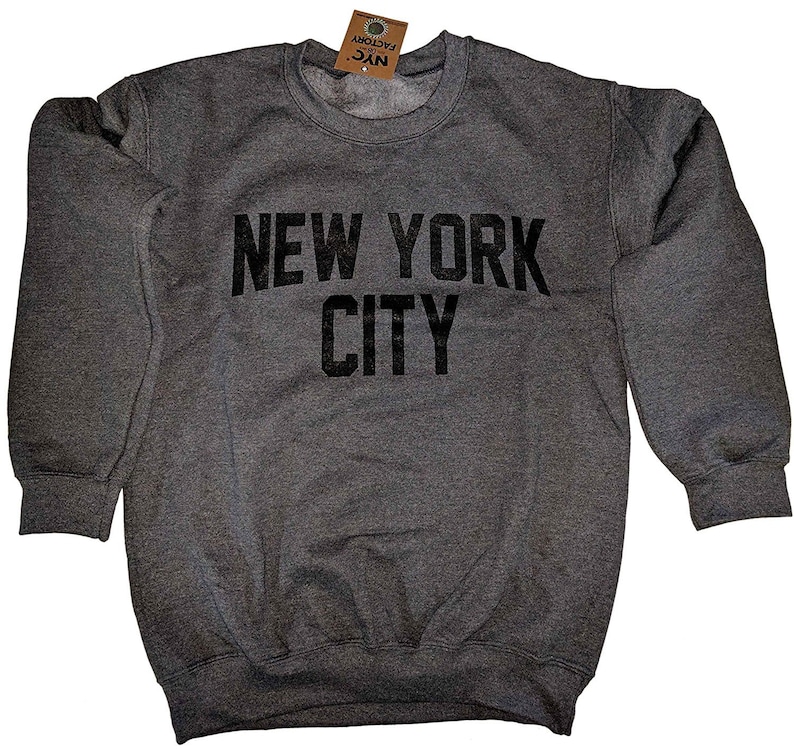 May include: A dark gray sweatshirt with the words "NEW YORK CITY" printed in large, black, block letters across the chest. The sweatshirt has a crew neck and long sleeves. A small tag is attached to the collar.