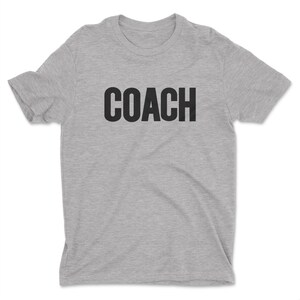 K&ouml;nnte beinhalten: Ein graues T-Shirt mit dem Wort "COACH" in Schwarz auf der Vorderseite.