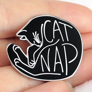 Può includere: Spilla in smalto nero con un contorno bianco di un gatto che dorme e il testo "CAT NAP".