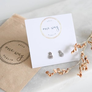 Könnte beinhalten: Ein Paar silberne Ananas-Ohrstecker auf einer weißen Karte mit dem Text "one way 2017 DUBLIN" in einem Kreis. Die Karte liegt auf einem braunen Papierbeutel mit dem gleichen Text.