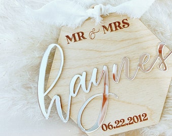 May include: A hexagonal wooden sign with the words "MR & MRS Haynes" and the date "06.22.2012" engraved in brown. The sign has a silver mirror-like finish and is decorated with a white ribbon.