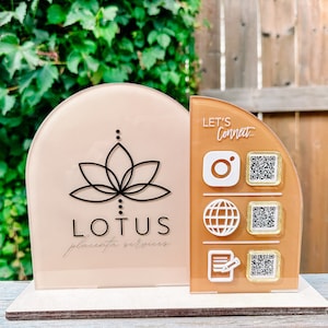 Peut inclure: Une enseigne en acrylique brun clair avec un logo de fleur de lotus et le texte "LOTUS placenta services". L'enseigne est &agrave; c&ocirc;t&eacute; d'une enseigne en acrylique brun clair avec le texte "LET'S Connect..." et trois ic&ocirc;nes avec des codes QR : Instagram, site web et email.