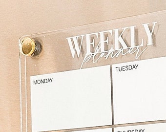 May include: A clear acrylic weekly planner with gold accents. The planner has spaces for each day of the week, Monday through Sunday, and a notes section.