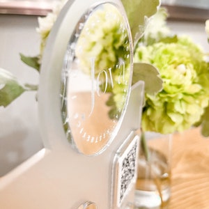 May include: A clear acrylic sign with the word "Jova" etched into it. The sign is attached to a white surface with a clear acrylic circle and a QR code.